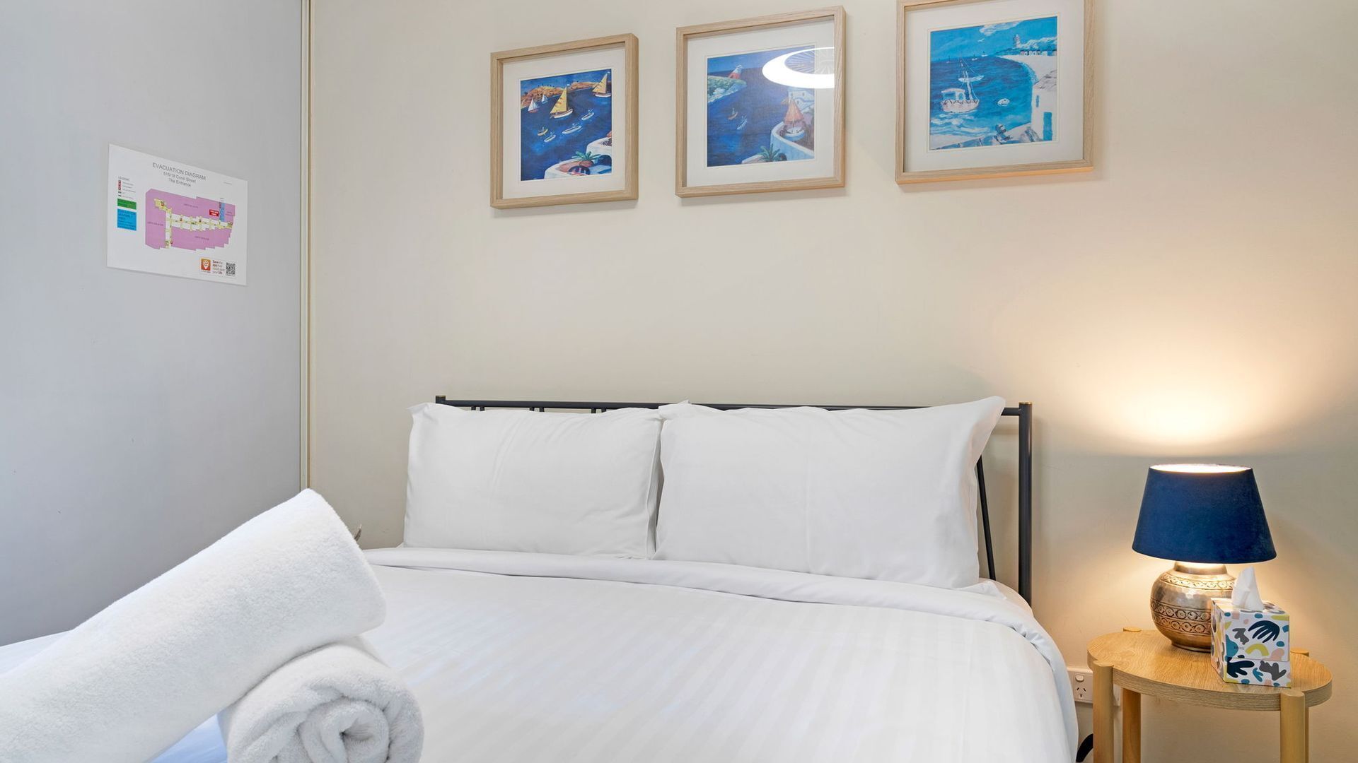 A hotel room with a bed, a nightstand with a lamp, and three framed artworks on the wall.