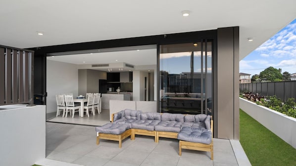 An open-plan living area with a dining table and chairs, a sofa, and a kitchen visible through a glass door. - The Accessible Oasis - 3 bedroom disability apartment (The Entrance)