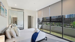 A modern bedroom with a large bed, a bedside table, and a window with a view of a courtyard. - The Accessible Oasis - 3 bedroom disability apartment (The Entrance)