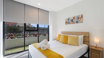 Habitación de hotel con una cama grande, una mesita de noche con lámpara y vistas a un jardín cercado a través de una puerta corredera de vidrio.