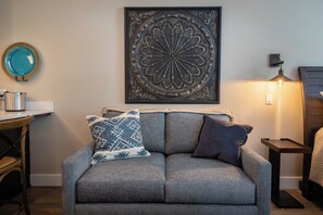 Een woonkamer met een grijze bank, een decoratief wandobject, een tafeltje met een waterkoker en een lamp.