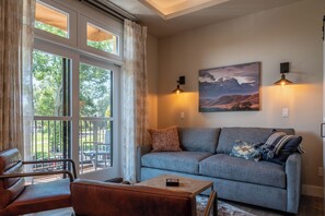 Ein gemütliches Wohnzimmer mit einem grauen Sofa, einem Ledersessel und einem kleinen Tisch. Zwei Wandlampen und ein Gemälde befinden sich über dem Sofa.