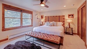 A bedroom with a large bed, a ceiling fan, a window with a view of trees, a bench, and a nightstand with a lamp.