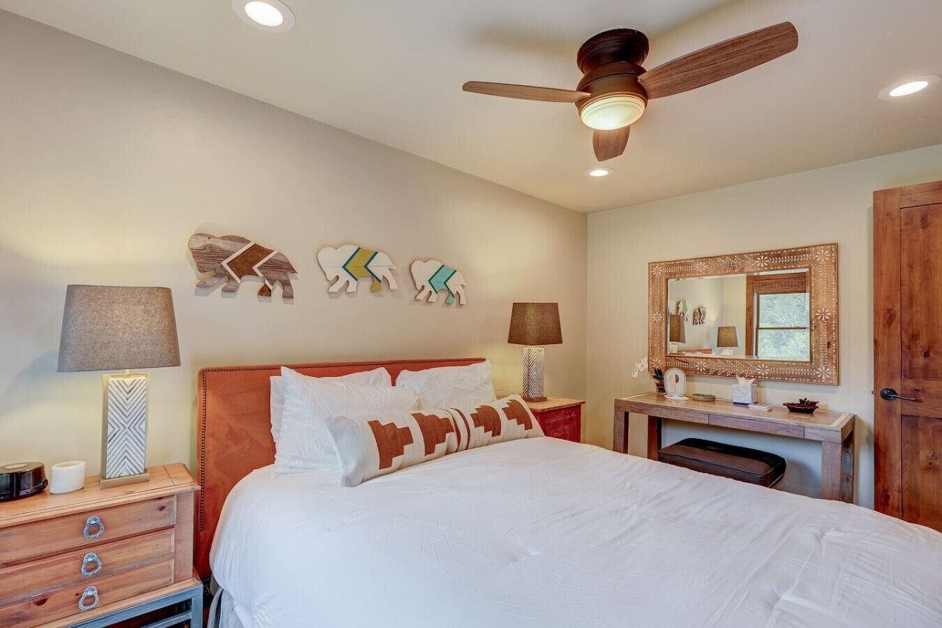 Quarto com cama, duas mesas de cabeceira, ventilador de teto, decoração na parede e um espelho.