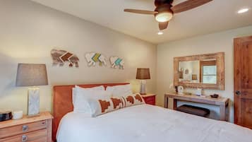 Quarto com cama, duas mesas de cabeceira, ventilador de teto, decoração na parede e um espelho.