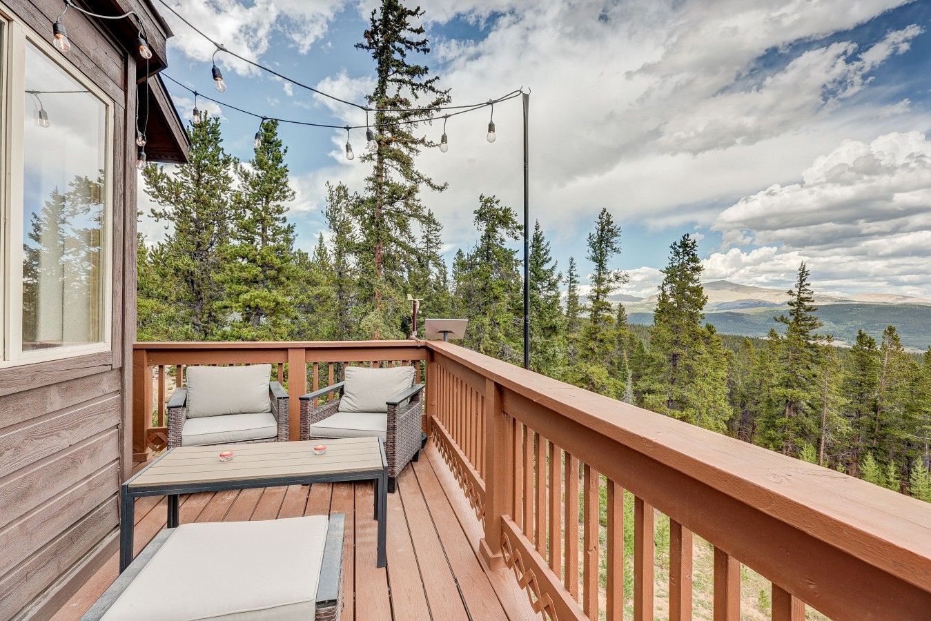 A wooden deck with outdoor seating, a view of a forest, and a mountain range in the distance.