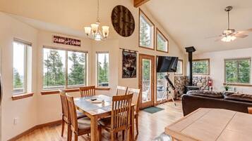 A spacious living area with a dining table, chairs, a fireplace, a television, and large windows offering views of the outdoors.