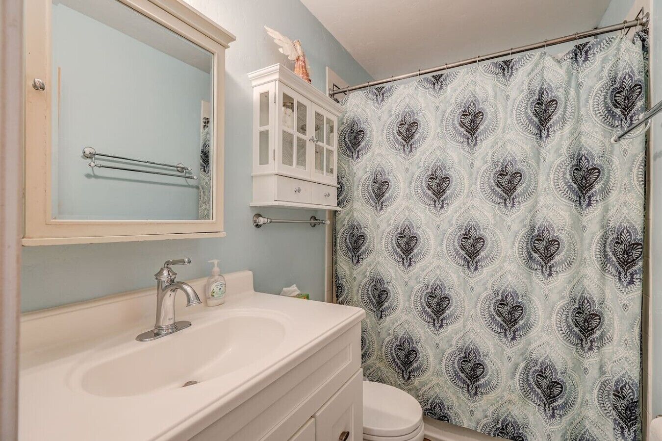 Un baño con tocador blanco, espejo, cortina de ducha con estampado de corazón y inodoro blanco.