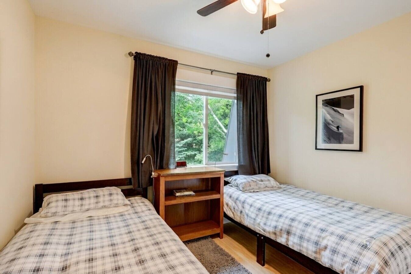 Une chambre avec deux lits, une fenêtre avec des rideaux, un ventilateur de plafond, un tableau encadré au mur et une petite table en bois.