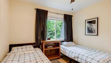 Une chambre avec deux lits, une fenêtre avec des rideaux, un ventilateur de plafond, un tableau encadré au mur et une petite table en bois.
