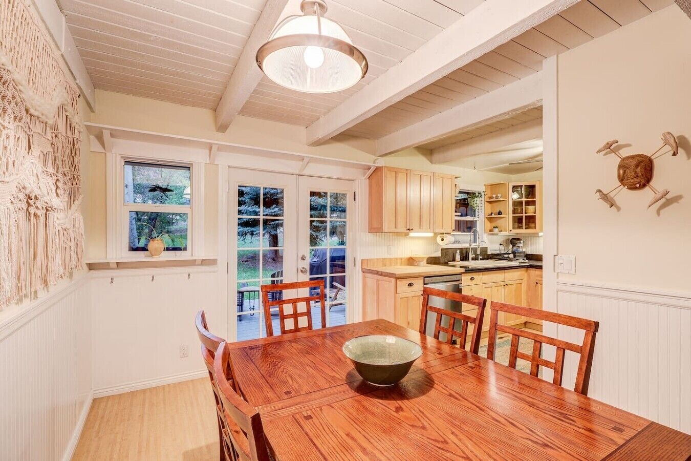 Un comedor con mesa y sillas de madera, una lámpara colgante y vista a la cocina a través de puertas de vidrio.