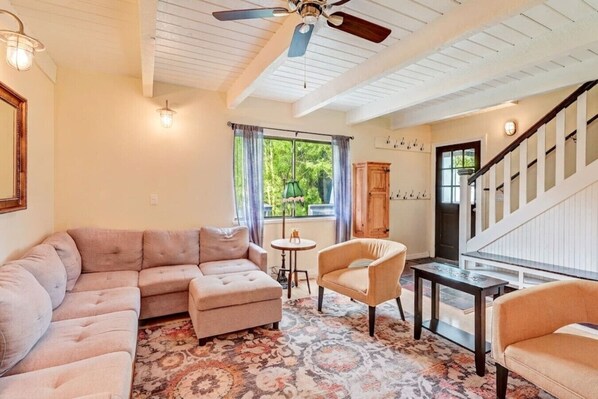 A living room with a sectional sofa, a round table, and a chair. There is a staircase with a railing and a door with a coat rack.