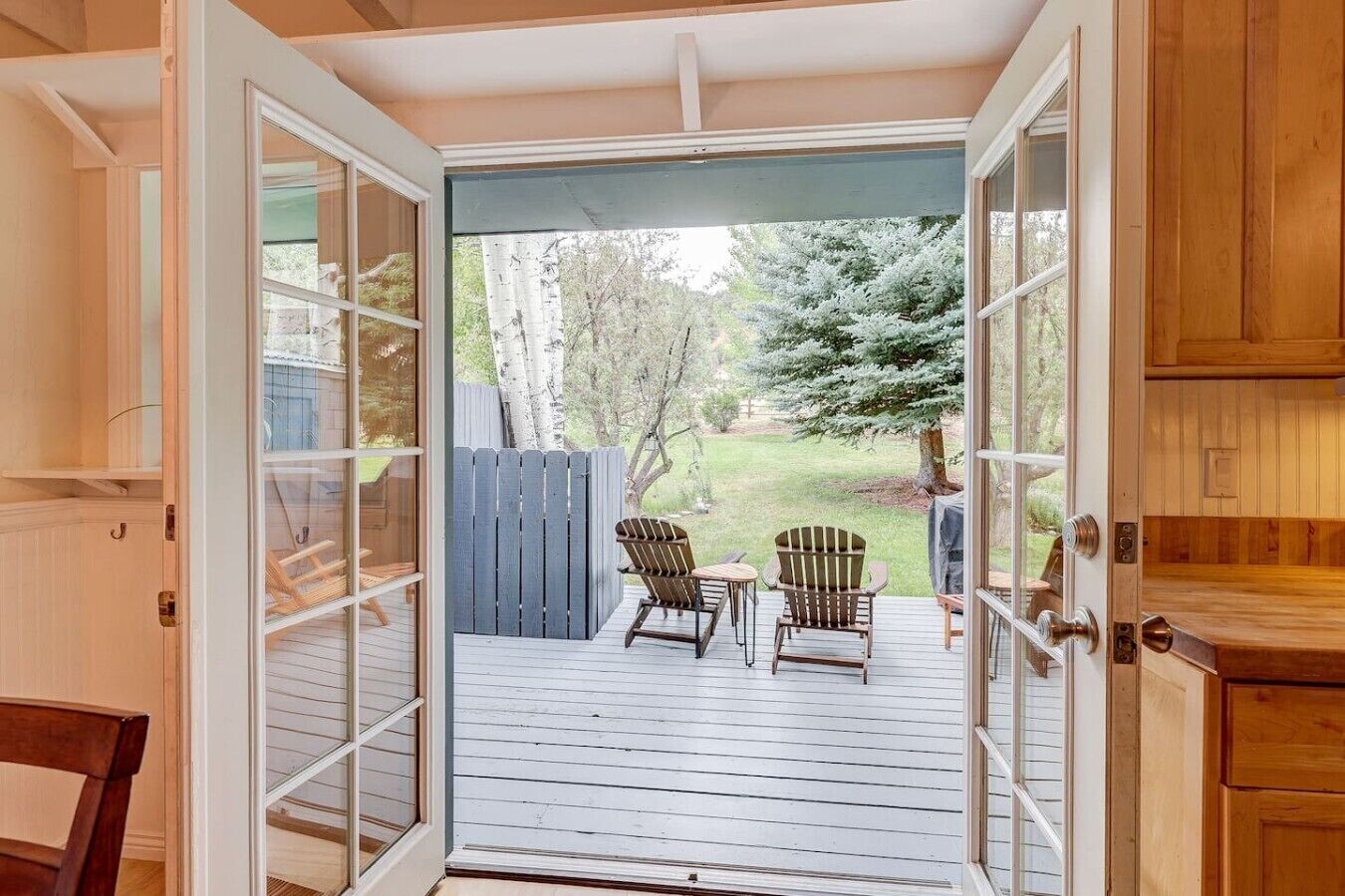 Une porte ouverte donne sur une terrasse où se trouvent deux chaises et une table, le tout étant entouré d’une clôture en bois et d’arbres.