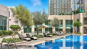A swimming pool with lounge chairs and umbrellas, surrounded by buildings and trees. - Burj Views Studio - Downtown Dubai -Bills Included (Dubai)