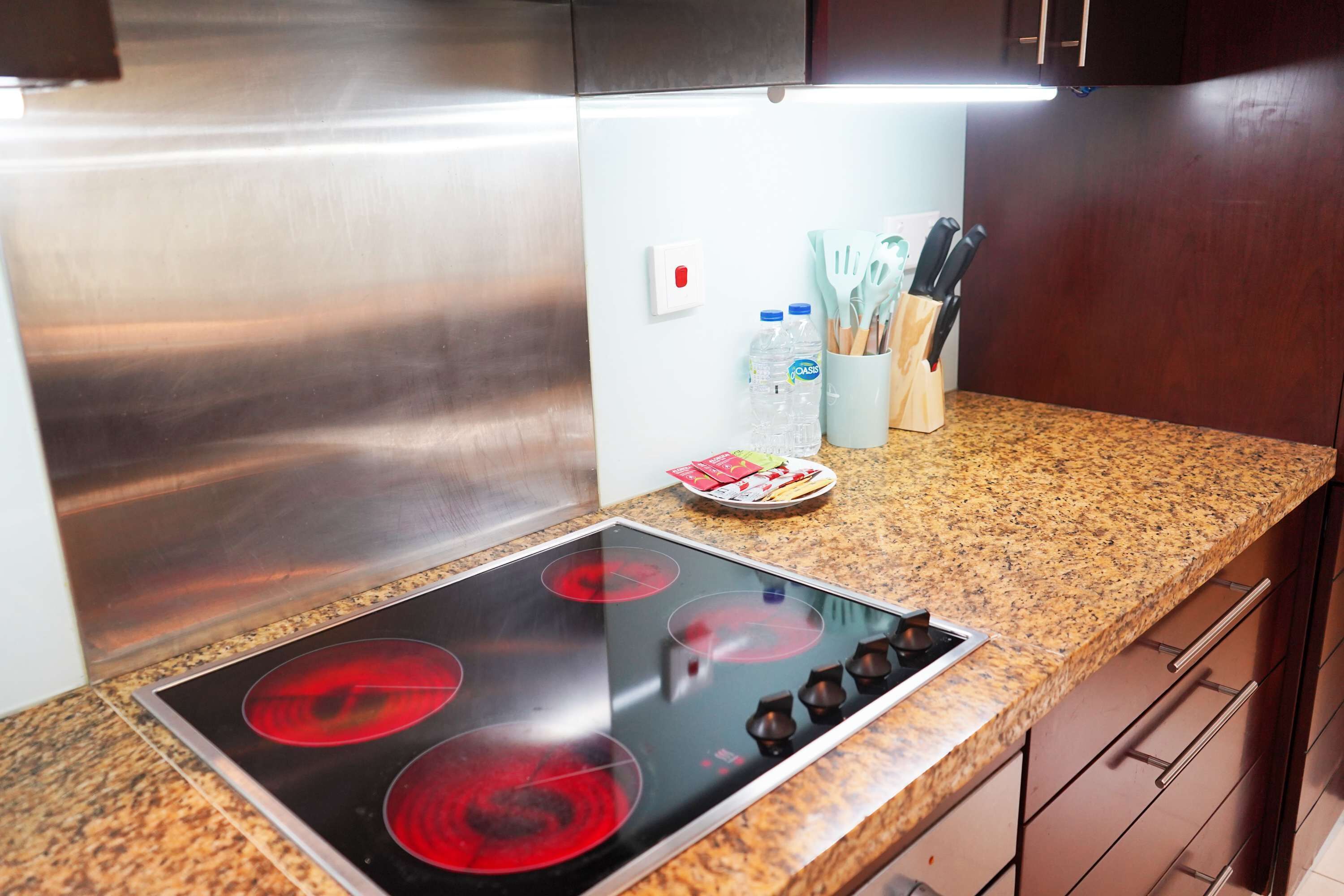 A kitchen with a stainless steel refrigerator, a granite countertop, a built-in stove with four burners, and a white wall with a power outlet.
