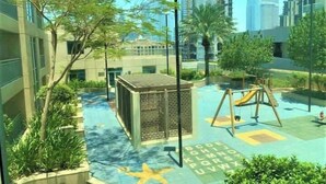 A children's play area with a slide and climbing structure, surrounded by greenery and modern buildings. - Burj Views Studio - Downtown Dubai -Bills Included (Dubai)