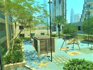 A children's play area with a slide and climbing structure, surrounded by greenery and modern buildings. - Burj Views Studio - Downtown Dubai -Bills Included (Dubai)