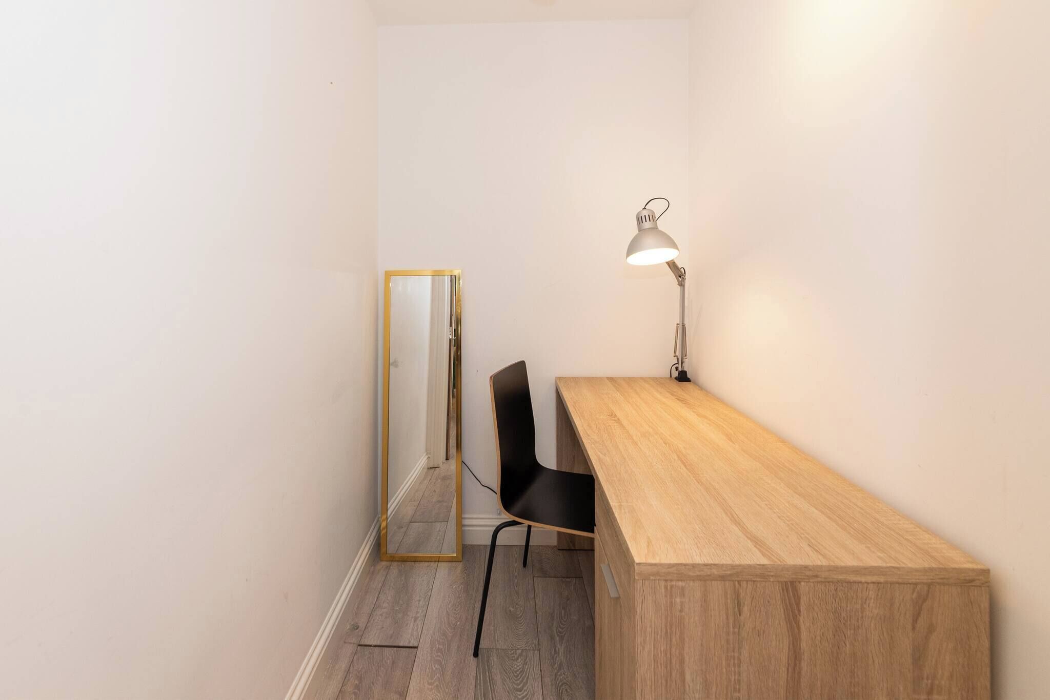 A small, narrow room with a wooden desk, a black chair, a floor lamp, and a mirror on the wall.