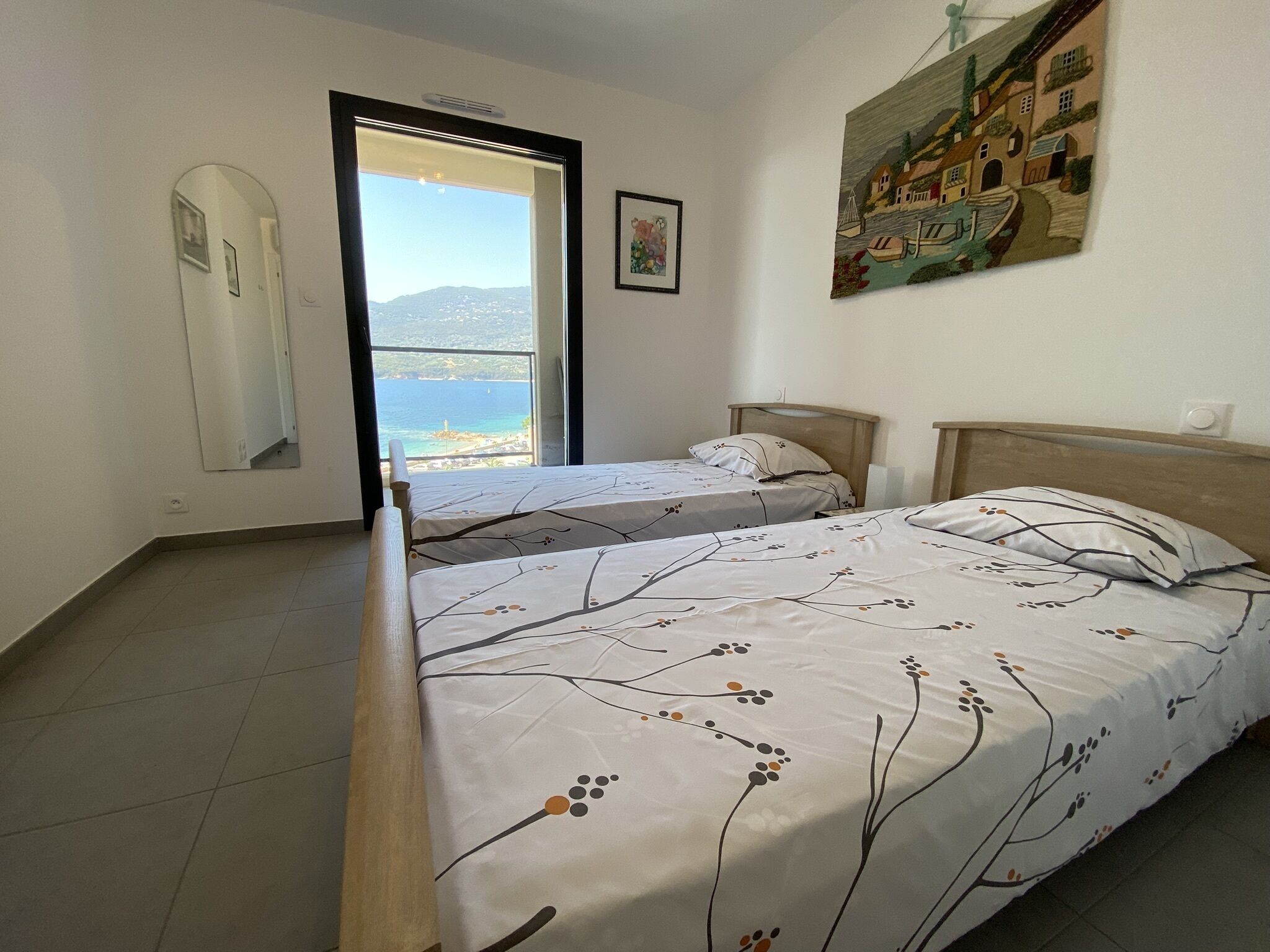 A bedroom with a large bed, a smaller bed, and a door leading to a balcony with a view of the sea.