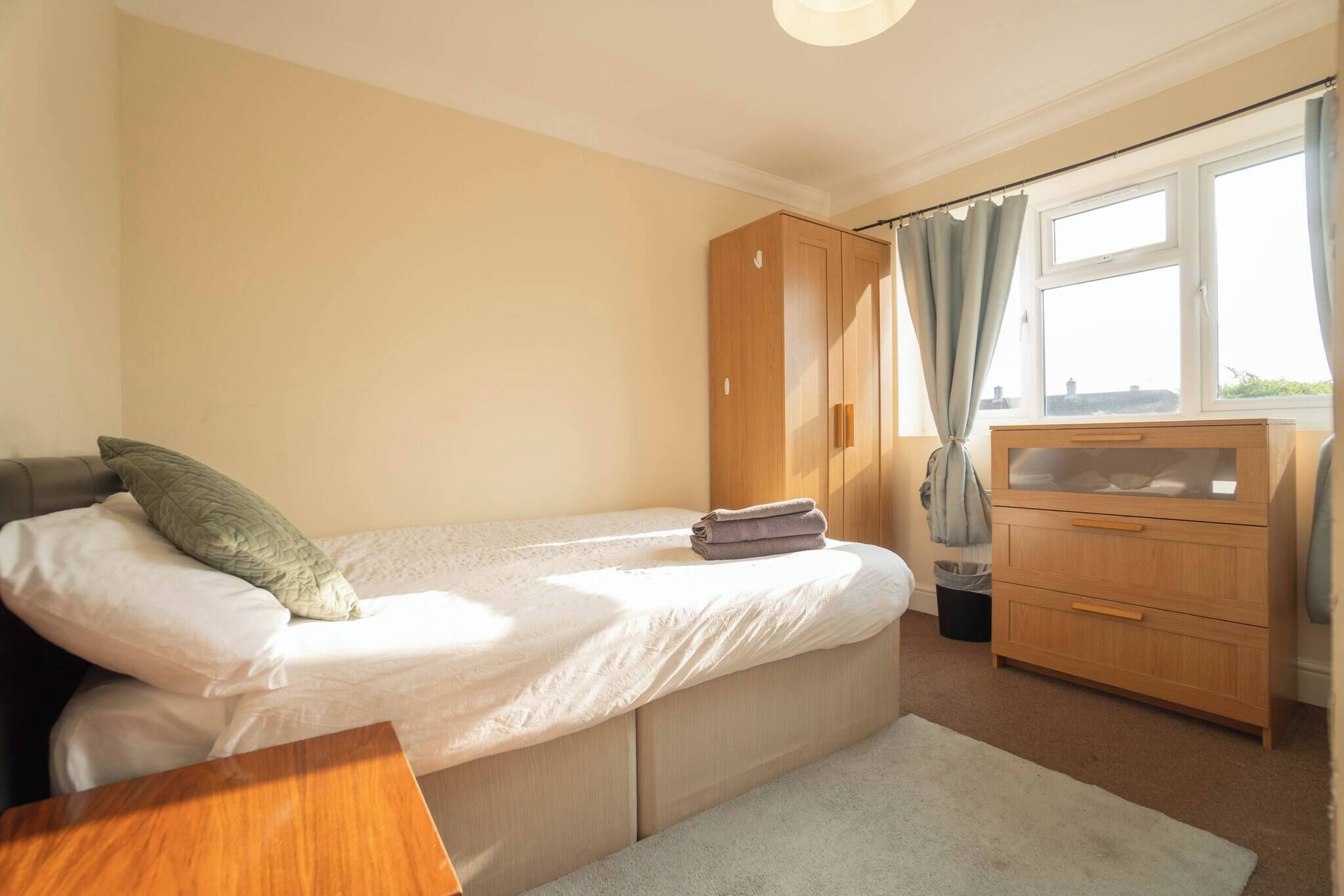 A bedroom with a single bed, a wooden wardrobe, a small desk, and a window with curtains.