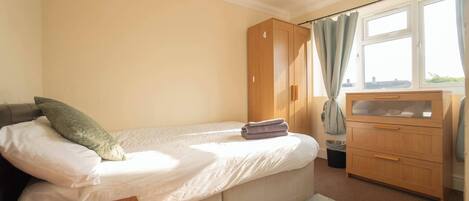A bedroom with a single bed, a wooden wardrobe, a small desk, and a window with curtains.