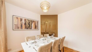 Un coin repas avec une table blanche dressée pour six personnes, comprenant des assiettes, des verres et des couverts. Un tableau abstrait encadré orne le mur, ainsi qu’un panneau en bois vertical.