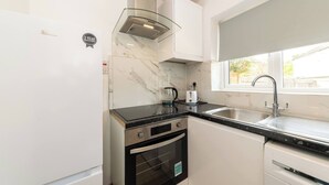 Une cuisine compacte avec des meubles blancs, un évier en acier inoxydable, une bouilloire en verre et un four encastré.