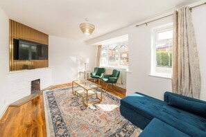 Ein Wohnzimmer mit blauem Sofa, einem gemusterten Teppich, einem an der Wand montierten Fernseher und einem Fenster mit Vorhängen.