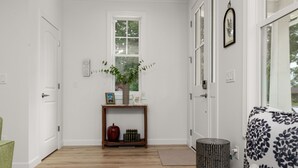 En hall med trägolv, en vit dörr, ett fönster med en växt, ett litet bord med en vas och en mönstrad soffa.