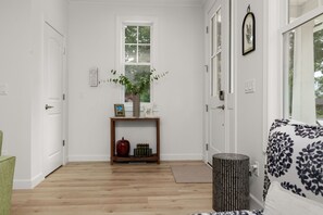 Ein Flur mit Holzboden, einer weißen Tür, einem Fenster mit Pflanze, einem kleinen Tisch mit Vase und einem gemusterten Sofa.