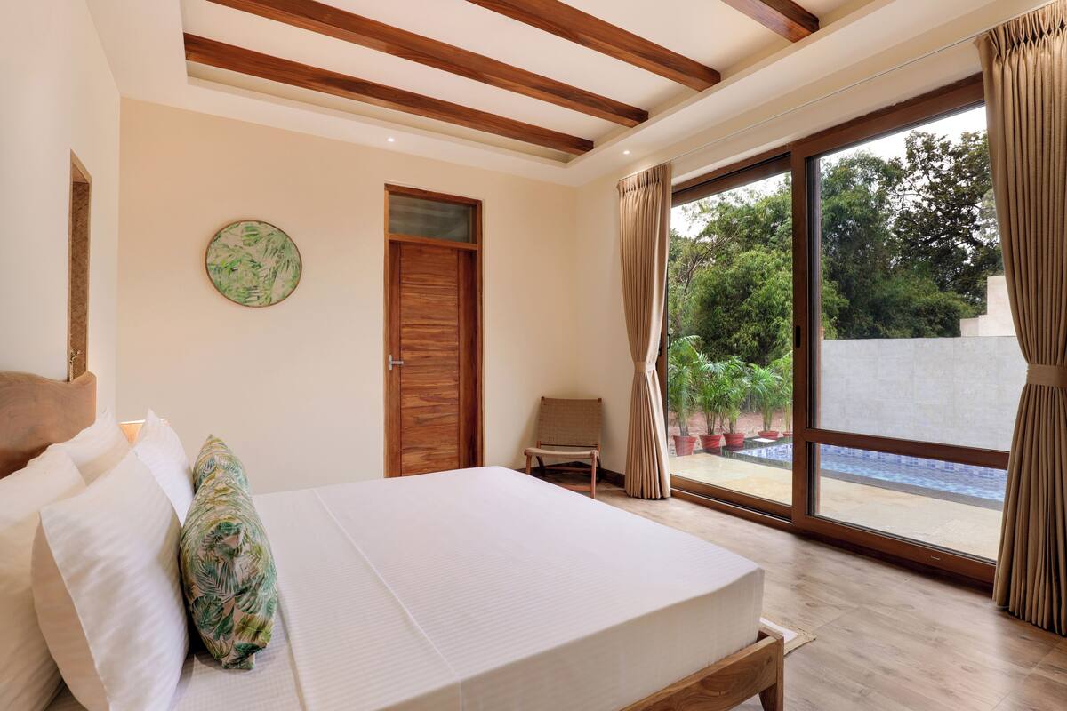 A bedroom with a bed, a wooden door, a chair, and a view of a pool through large windows.
