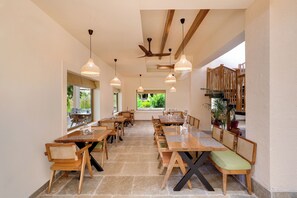 A spacious dining area with wooden tables and chairs, pendant lighting, and a view of greenery outside. - The Safari Lodge Kanha  (Baihar)