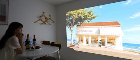A room with a large projection screen displaying a coastal view, a dining table with chairs, and a few bottles and glasses.