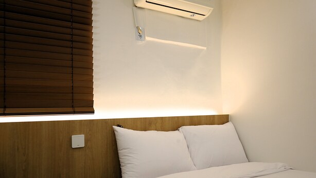 A hotel room with a wooden headboard, a bed with white bedding, a wall-mounted air conditioning unit, and a light switch.