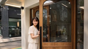 A person standing at a wooden door with glass panels, a street visible outside, and a building with signage in the background.