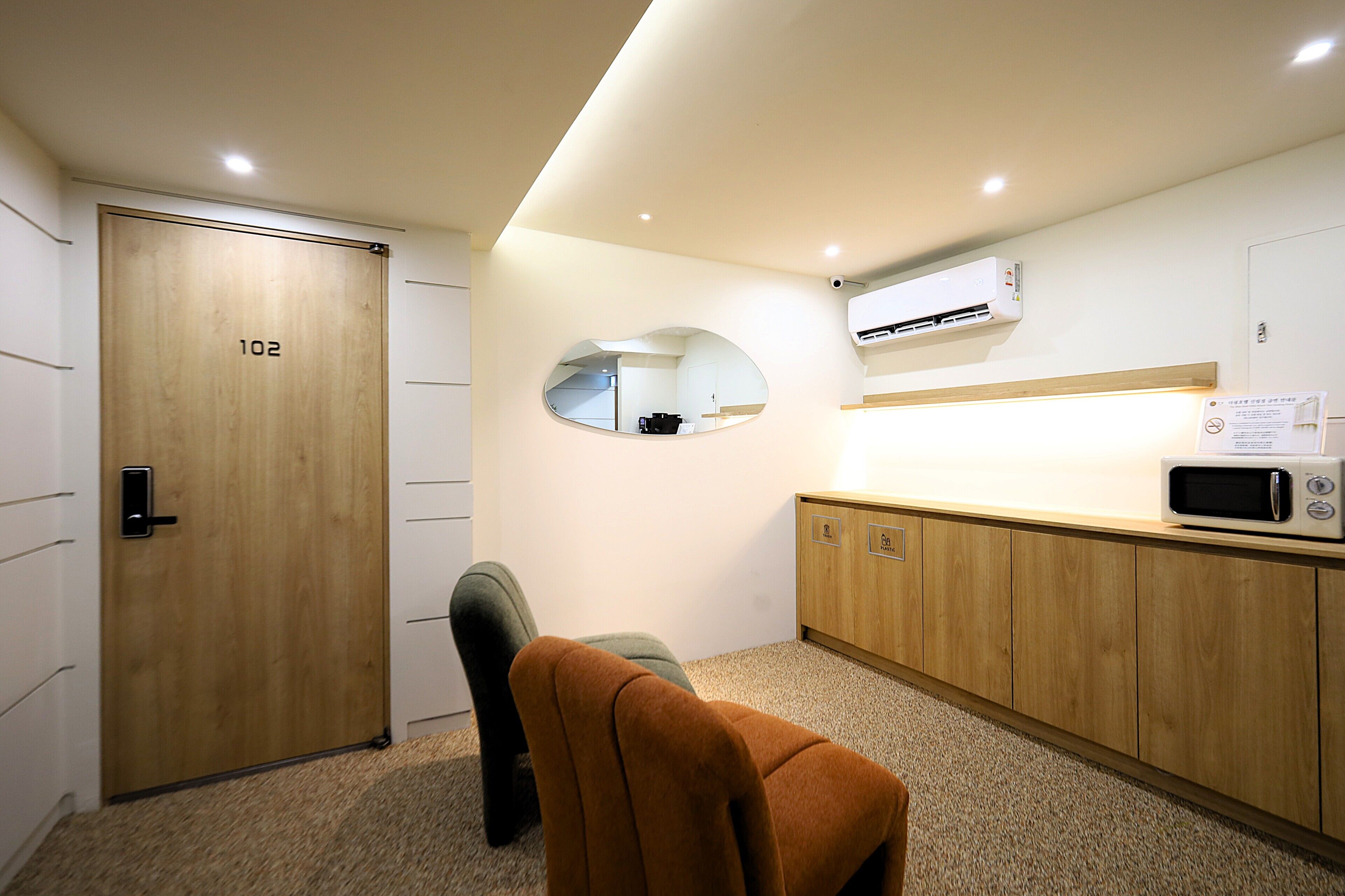 Hotelkamer met een houten deur waarop '102' staat, een klein rond spiegel, een magnetron en een stoel met een plaid.