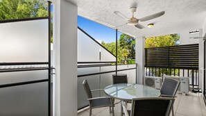 A balcony with a glass table and chairs, a ceiling fan, and air conditioning units. - Palm Cove One-Bedroom Apartment with Pool | Oliva Breeze Apartment (Palm Cove)