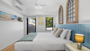 A bedroom with a bed, a nightstand, a lamp, and a window with a view of greenery. - Palm Cove One-Bedroom Apartment with Pool | Oliva Breeze Apartment (Palm Cove)