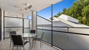 A modern outdoor balcony with a glass table and chairs, a ceiling fan, and a view of a house. - Palm Cove One-Bedroom Apartment with Pool | Myrtle at Oliva (Palm Cove)