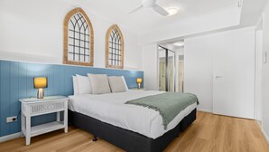 Quarto com cama, mesas de cabeceira, ventilador de teto e porta que dá para outro espaço.