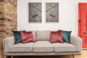 A neutral-colored sofa with three cushions against a white wall, flanked by a brick wall on one side and a red door on the other. - Newly renovated 2 Bdrm Terrace + Rooftop (City of Sydney)