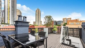 Eine Dachterrasse mit Glastisch und Stühlen, umgeben von einer Holzterrasse und Blick auf die Stadt.