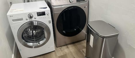 Een wasruimte met een voorlader wasmachine en droger, een afvalbak van roestvrij staal en een houten vloer.