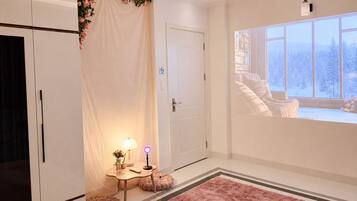 A bedroom with a bed, a bedside table, a lamp, a window with a view of snow-covered trees, and a door with a no-smoking sign.