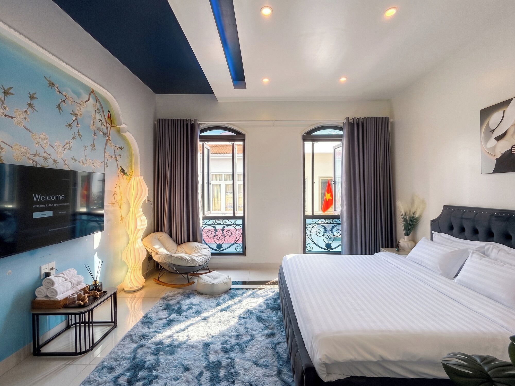 A modern hotel room with a large bed, a seating area with a chair, a TV mounted on the wall, and a decorative wall piece.