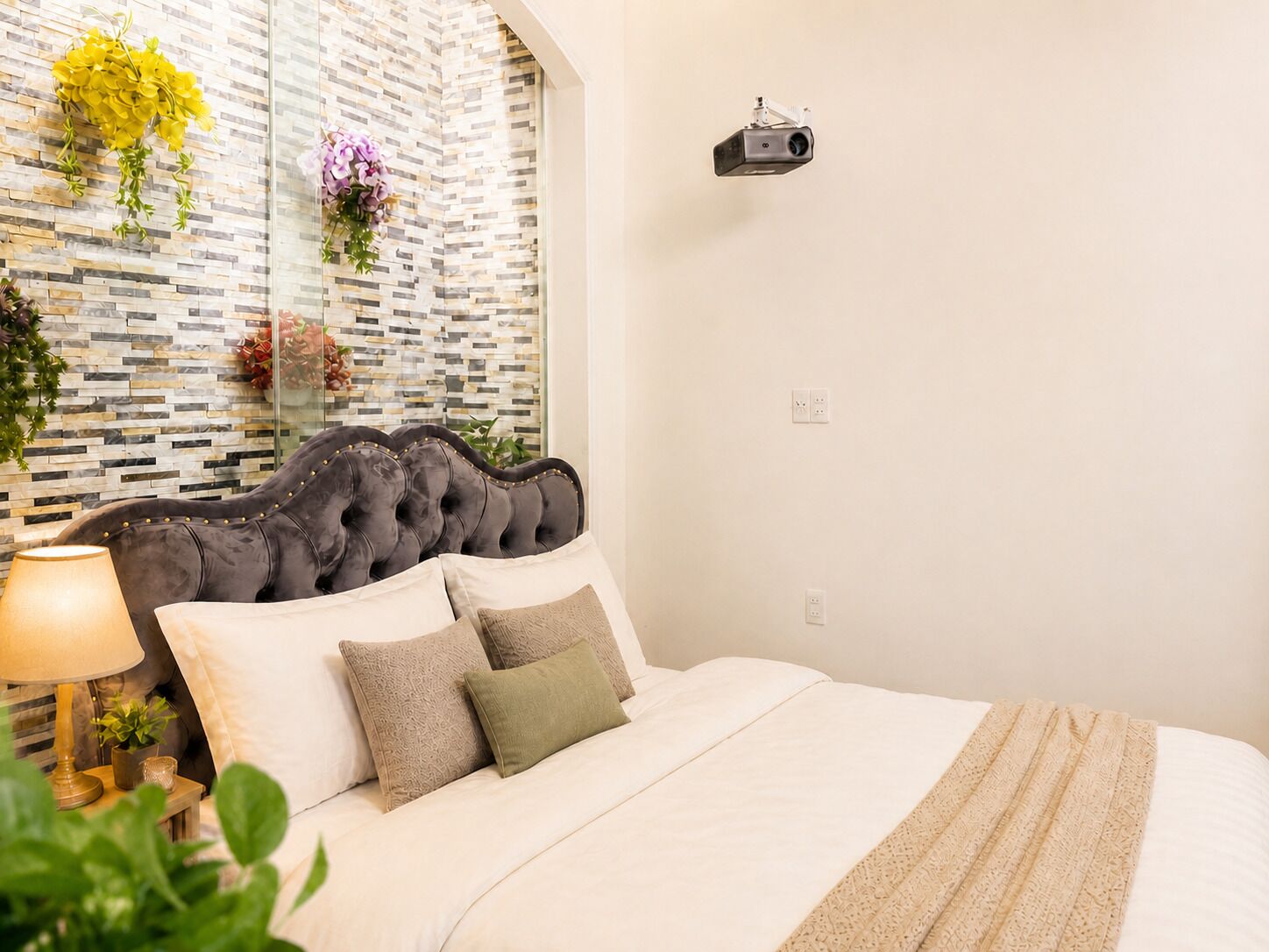 A bedroom with a tufted headboard, a bed with white linens, a lamp, and decorative plants.