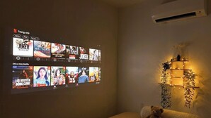 Un dormitorio con una televisión de montaje en pared mostrando diferentes programas, una cama con un osito de peluche y una estantería con objetos decorativos.