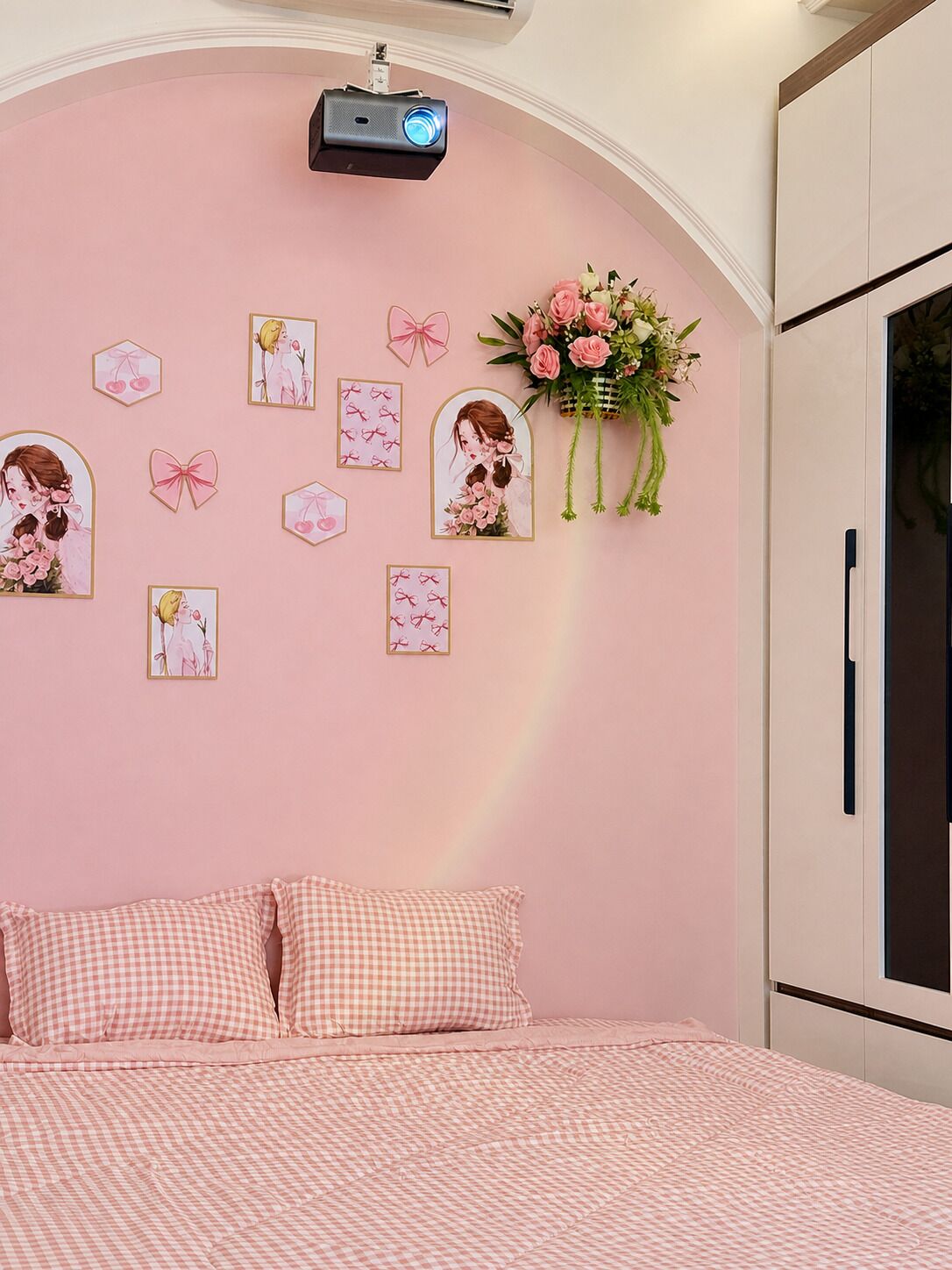 Een slaapkamer met een roze wand, een projector aan het plafond, een bed met geruit beddengoed, en wanddecoraties waaronder ingelijste foto’s en een bloemenboeket.