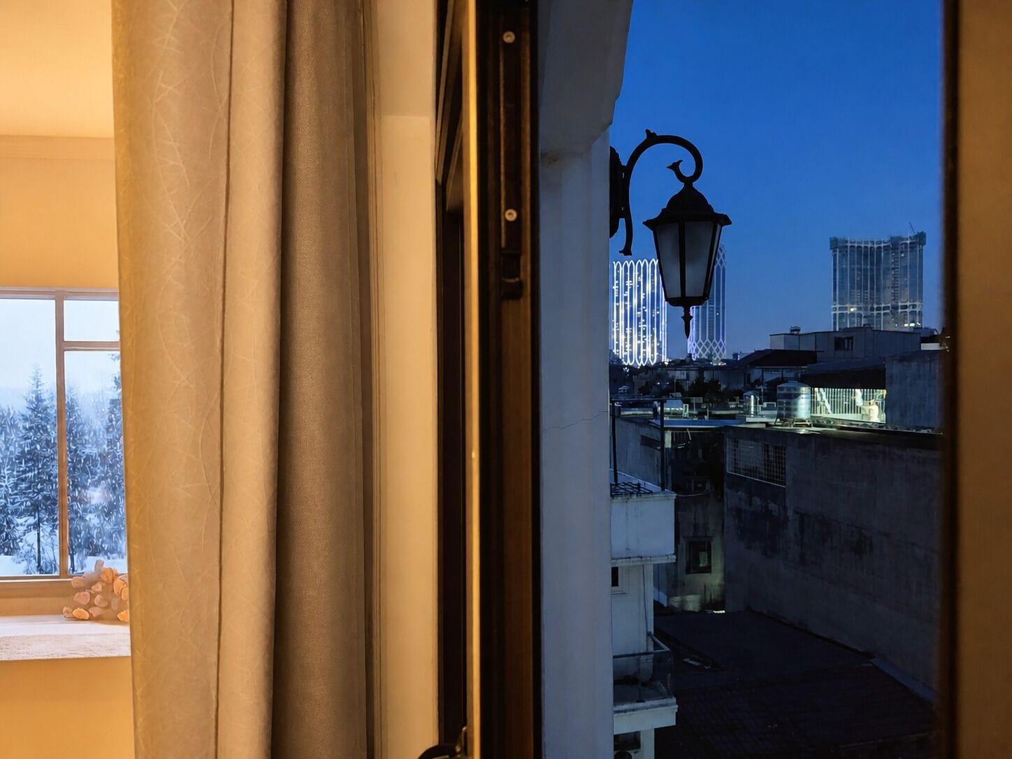 A view through a window with curtains, featuring a cityscape with buildings and a street lamp.