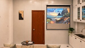 Una sala de estar con un sofá, una pequeña mesa de centro, una lámpara de araña, un cuadro y una pantalla de televisión grande que muestra un paisaje.